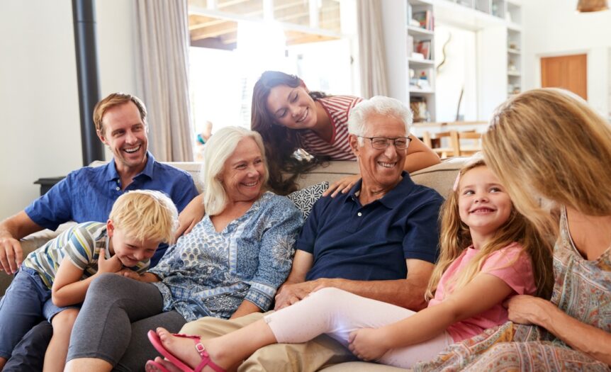 Two young children smile as they sit on a couch with three middle aged adults and two elderly people.
