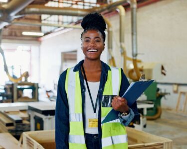 A young Black woman with a bright smile standing on a warehouse floor. She is wearing hi-vis gear and holding a clipboard.