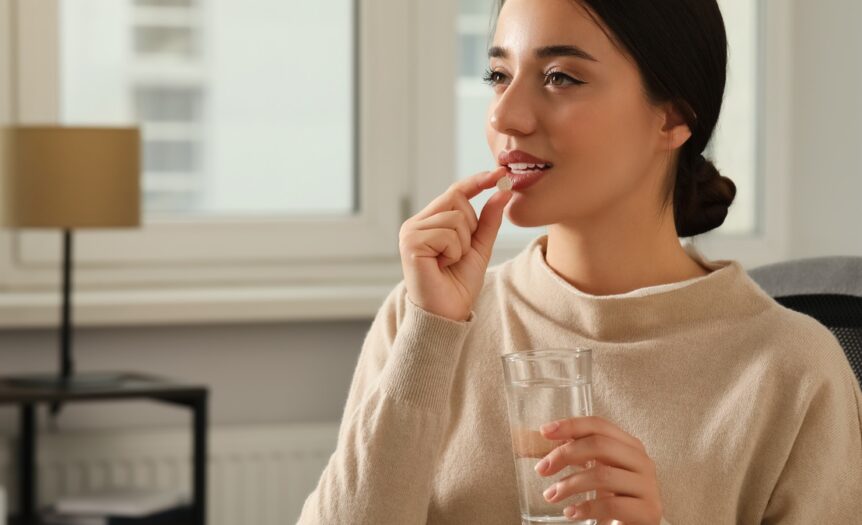 A woman with dark hair and a beige sweater puts a pill into her mouth while holding a glass of water.