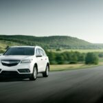 On a large road in the middle of nowhere, surrounded by hills and small trees, a small white car drives down the road.