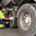 A trailer hauler kneeling on the ground next to their trailer's large tires. They are wearing neon yellow safety gear.