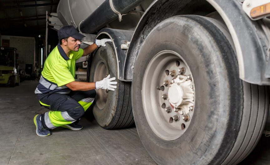A trailer hauler kneeling on the ground next to their trailer's large tires. They are wearing neon yellow safety gear.