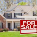 An in-focus red and white for sale sign in front of an out-of-focus home with a well-maintained lawn.
