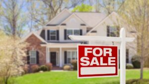 An in-focus red and white for sale sign in front of an out-of-focus home with a well-maintained lawn.