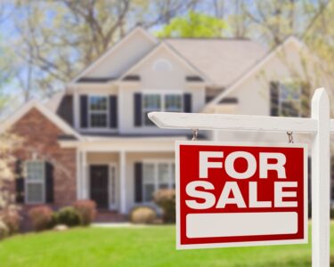 An in-focus red and white for sale sign in front of an out-of-focus home with a well-maintained lawn.