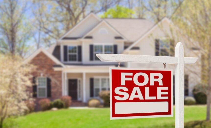 An in-focus red and white for sale sign in front of an out-of-focus home with a well-maintained lawn.