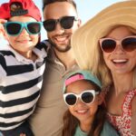 An adult man and woman standing on the beach with a young boy and girl. They're all smiling and wearing sunglasses.