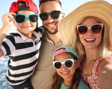 An adult man and woman standing on the beach with a young boy and girl. They're all smiling and wearing sunglasses.