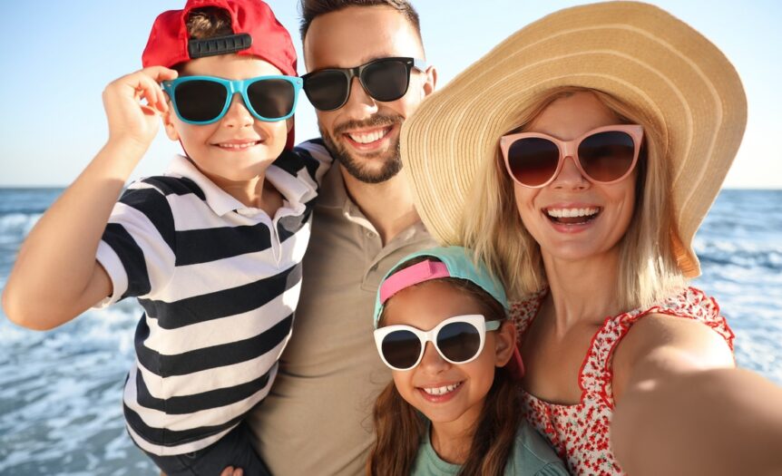 An adult man and woman standing on the beach with a young boy and girl. They're all smiling and wearing sunglasses.