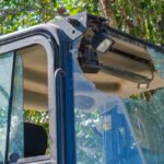 A broken windshield and a hanging light with exposed wiring are visible on an excavator parked near a tree in daylight.