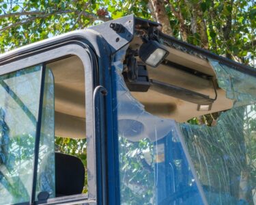A broken windshield and a hanging light with exposed wiring are visible on an excavator parked near a tree in daylight.