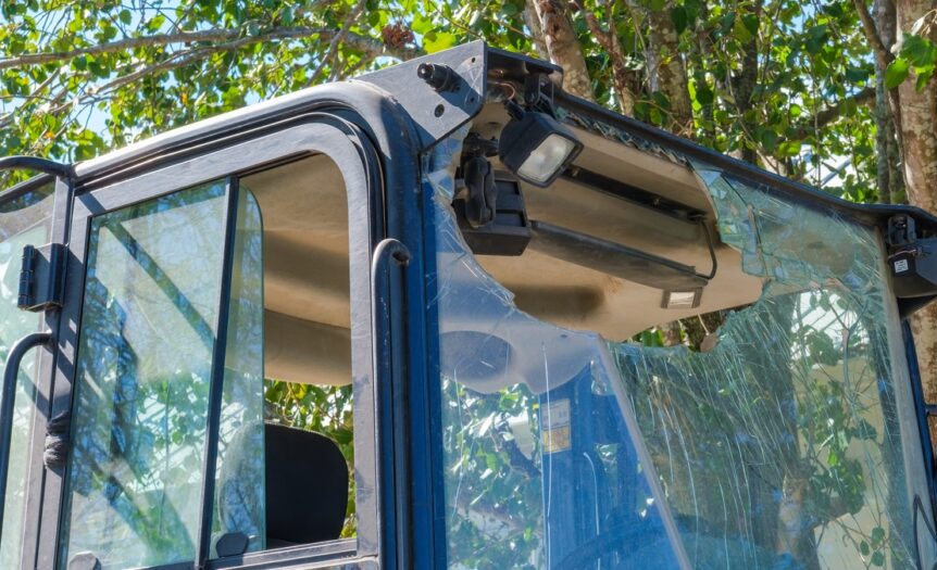A broken windshield and a hanging light with exposed wiring are visible on an excavator parked near a tree in daylight.