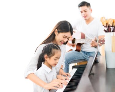 A woman helps her daughter at the piano while the father sits behind them and strums on an acoustic guitar.