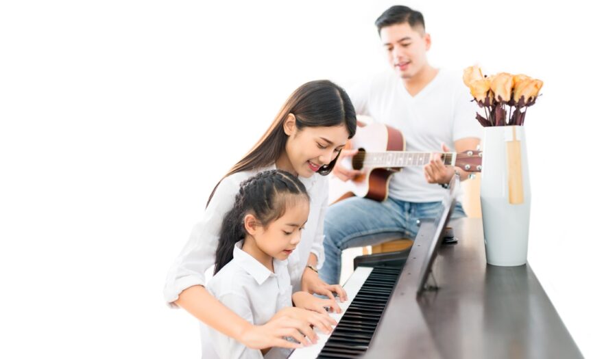 A woman helps her daughter at the piano while the father sits behind them and strums on an acoustic guitar.