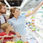 A family of three at the grocery store in the dairy section discussing milk types. The mom is holding a jug of milk.