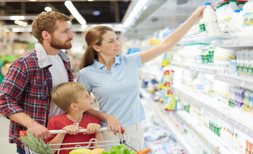 A family of three at the grocery store in the dairy section discussing milk types. The mom is holding a jug of milk.