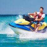 A bearded man and young boy wearing red and blue life vests ride a blue and yellow jet ski on a sunny day.