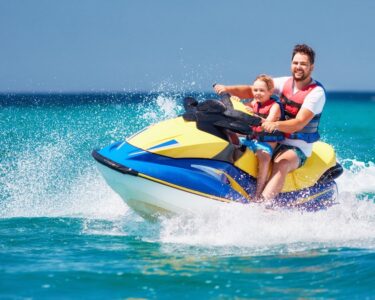 A bearded man and young boy wearing red and blue life vests ride a blue and yellow jet ski on a sunny day.