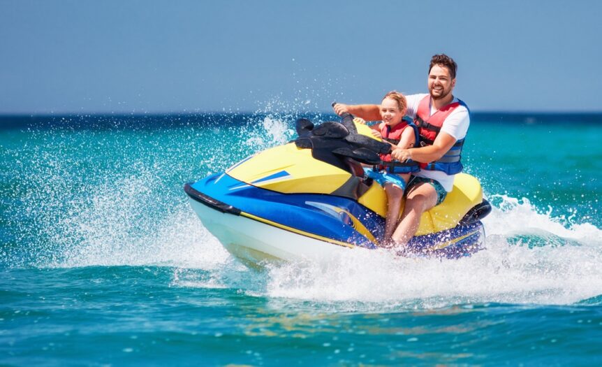 A bearded man and young boy wearing red and blue life vests ride a blue and yellow jet ski on a sunny day.