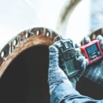 A person wearing stripped gloves holds two gas detection devices during an inspection up to a rusty vent.