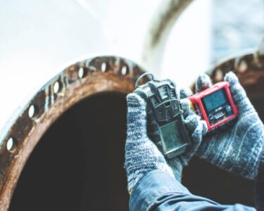 A person wearing stripped gloves holds two gas detection devices during an inspection up to a rusty vent.