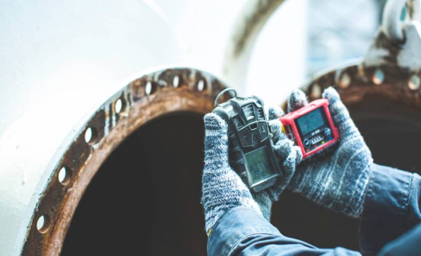 A person wearing stripped gloves holds two gas detection devices during an inspection up to a rusty vent.