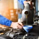 A close-up of a person wearing a blue jumpsuit and pouring oil into a blue funnel to replace a car engine’s oil.