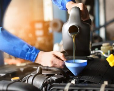 A close-up of a person wearing a blue jumpsuit and pouring oil into a blue funnel to replace a car engine’s oil.