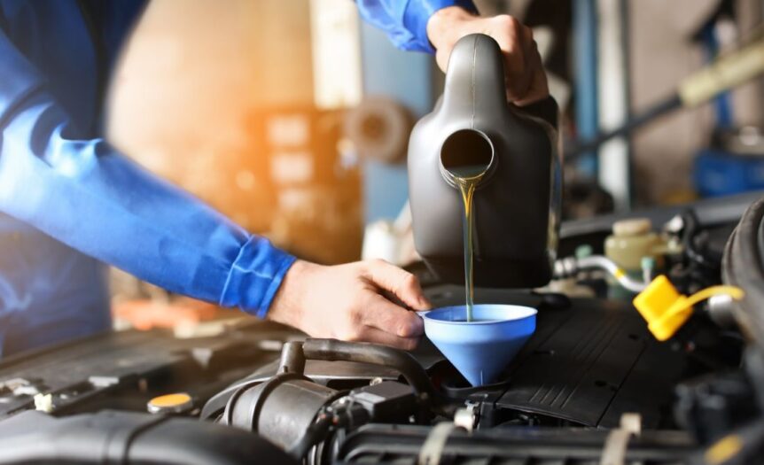 A close-up of a person wearing a blue jumpsuit and pouring oil into a blue funnel to replace a car engine’s oil.