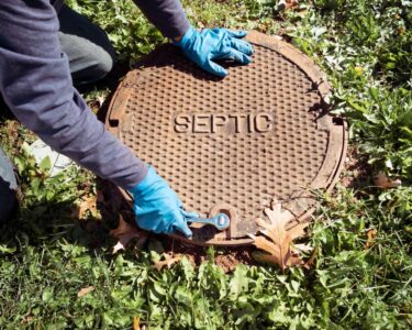 A worker wearing blue gloves holds a wrench to open up a septic tank opening in a homeowner's backyard.