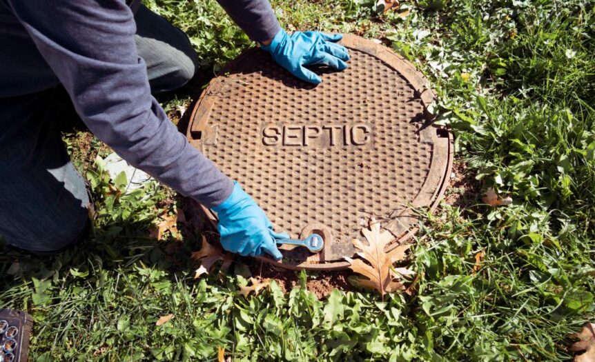 A worker wearing blue gloves holds a wrench to open up a septic tank opening in a homeowner's backyard.