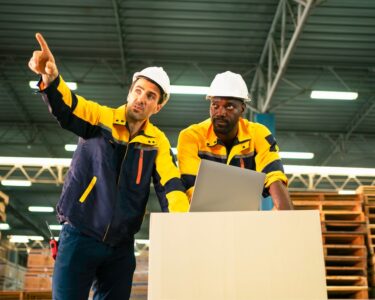 Two men in matching jackets and hard hats using a laptop inside a warehouse full of stacked wooden pallets.