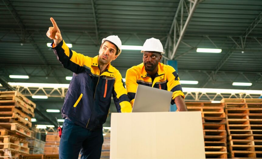 Two men in matching jackets and hard hats using a laptop inside a warehouse full of stacked wooden pallets.