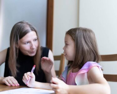 A little girl sits at a kitchen table with a pen in hand and schoolwork in front of her as her mother sits next to her and helps.
