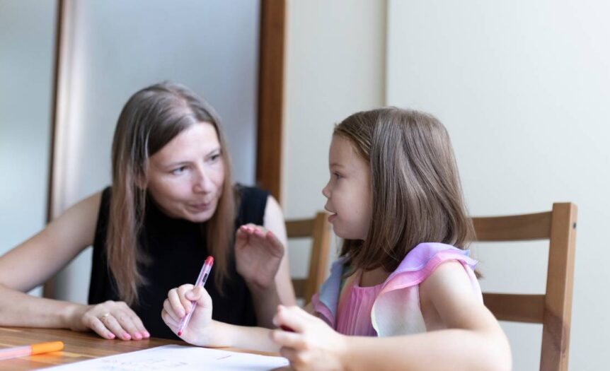 A little girl sits at a kitchen table with a pen in hand and schoolwork in front of her as her mother sits next to her and helps.