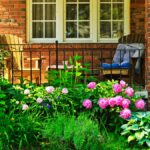 A close-up of a brick house's front porch with colorful flowers and two wooden chairs, one yellow and the other brown.