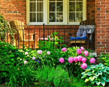 A close-up of a brick house's front porch with colorful flowers and two wooden chairs, one yellow and the other brown.