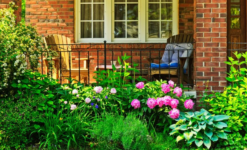 A close-up of a brick house's front porch with colorful flowers and two wooden chairs, one yellow and the other brown.