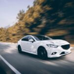 A shiny, white sedan drives quickly around a bend in the road. The surrounding scenery is blurred from movement.