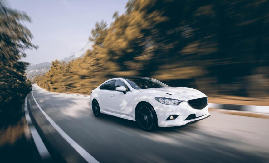 A shiny, white sedan drives quickly around a bend in the road. The surrounding scenery is blurred from movement.
