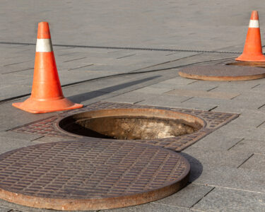 A manhole cover next to an opening in the sidewalk. There are two orange safety cones sitting next to the open hole.