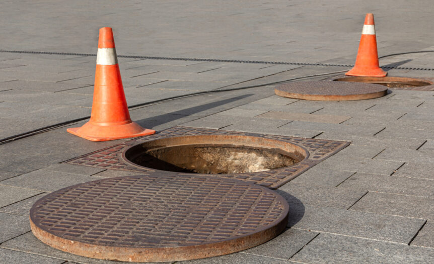 A manhole cover next to an opening in the sidewalk. There are two orange safety cones sitting next to the open hole.