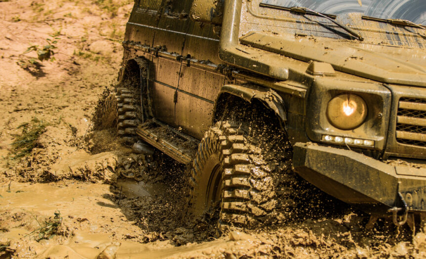 A Jeep’s headlights shine as mud splatters around the tires, and windshield wipers scrape thick mud away.