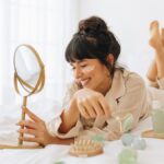 A woman with brown hair grins to herself. She lays on her bed, surrounded by different skin-care items.