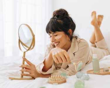 A woman with brown hair grins to herself. She lays on her bed, surrounded by different skin-care items.