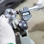 An operator wearing white gloves uses a digital micrometer to measure a newly manufactured circular silver metal part.