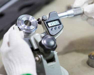 An operator wearing white gloves uses a digital micrometer to measure a newly manufactured circular silver metal part.