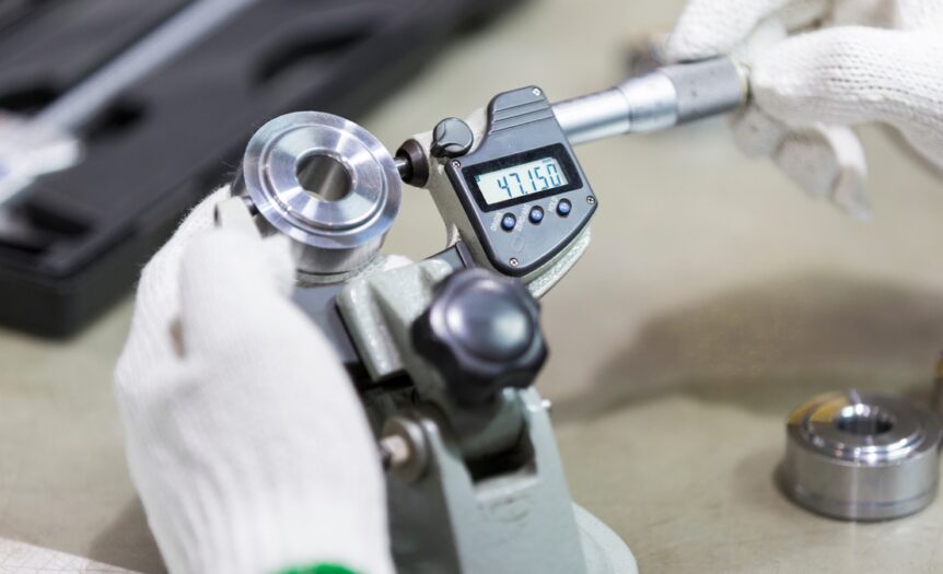 An operator wearing white gloves uses a digital micrometer to measure a newly manufactured circular silver metal part.