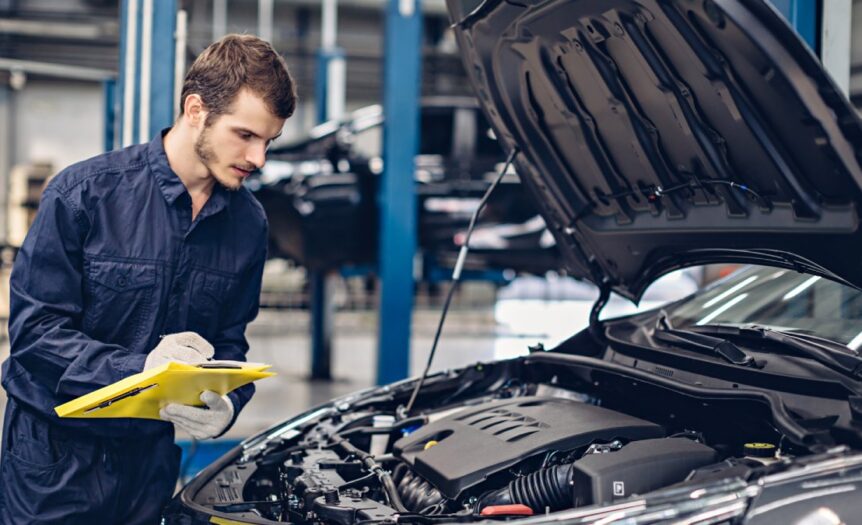 An auto mechanic in blue overalls examines the exposed engine bay of a black sedan and takes notes on a clipboard.