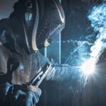 A man wearing a helmet and a fire-resistant bodysuit uses a welding torch on a piece of metal, sending out sparks and smoke.